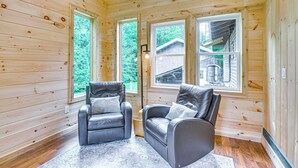 A wooden cabin interior with two leather chairs, large windows, and a visible wooden deck outside.