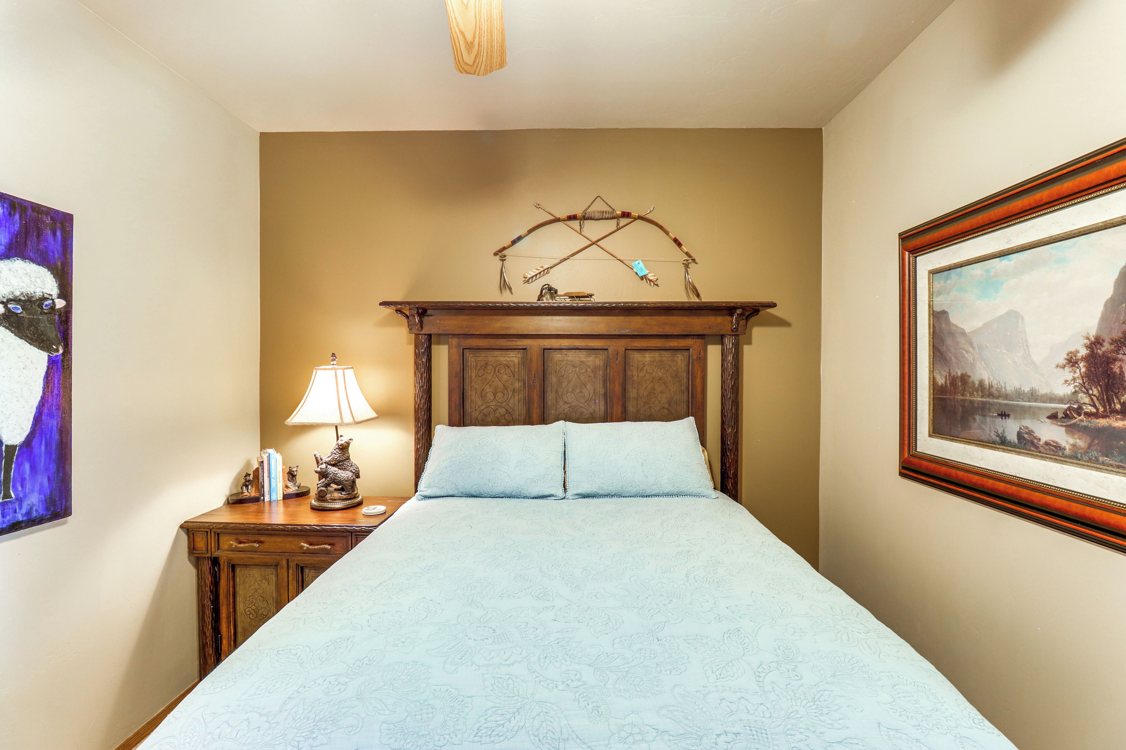 A bedroom with a wooden headboard, a bed with a patterned bedspread, a nightstand with a lamp and a small statue, and two framed artworks on the walls.