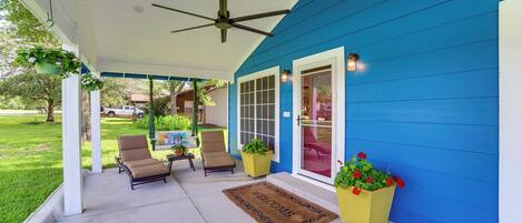A covered porch with a ceiling fan, two lounge chairs, a welcome mat, and potted plants.