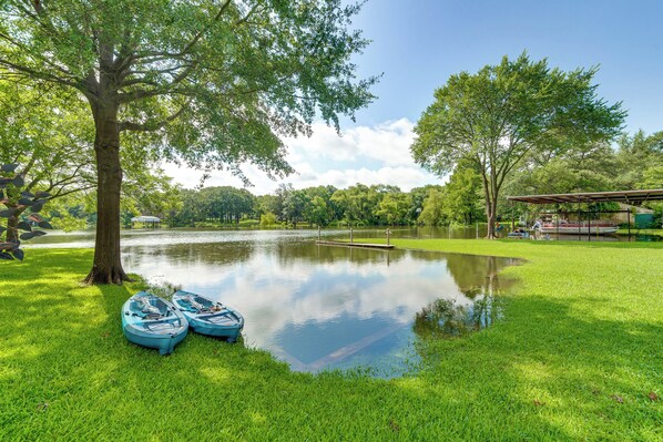 A serene lakeside setting with two kayaks docked near a tree, a grassy area, and a pavilion in the background. - Lake Tawakoni Access + Hot Tub - Wills Point Home! (Wills Point)