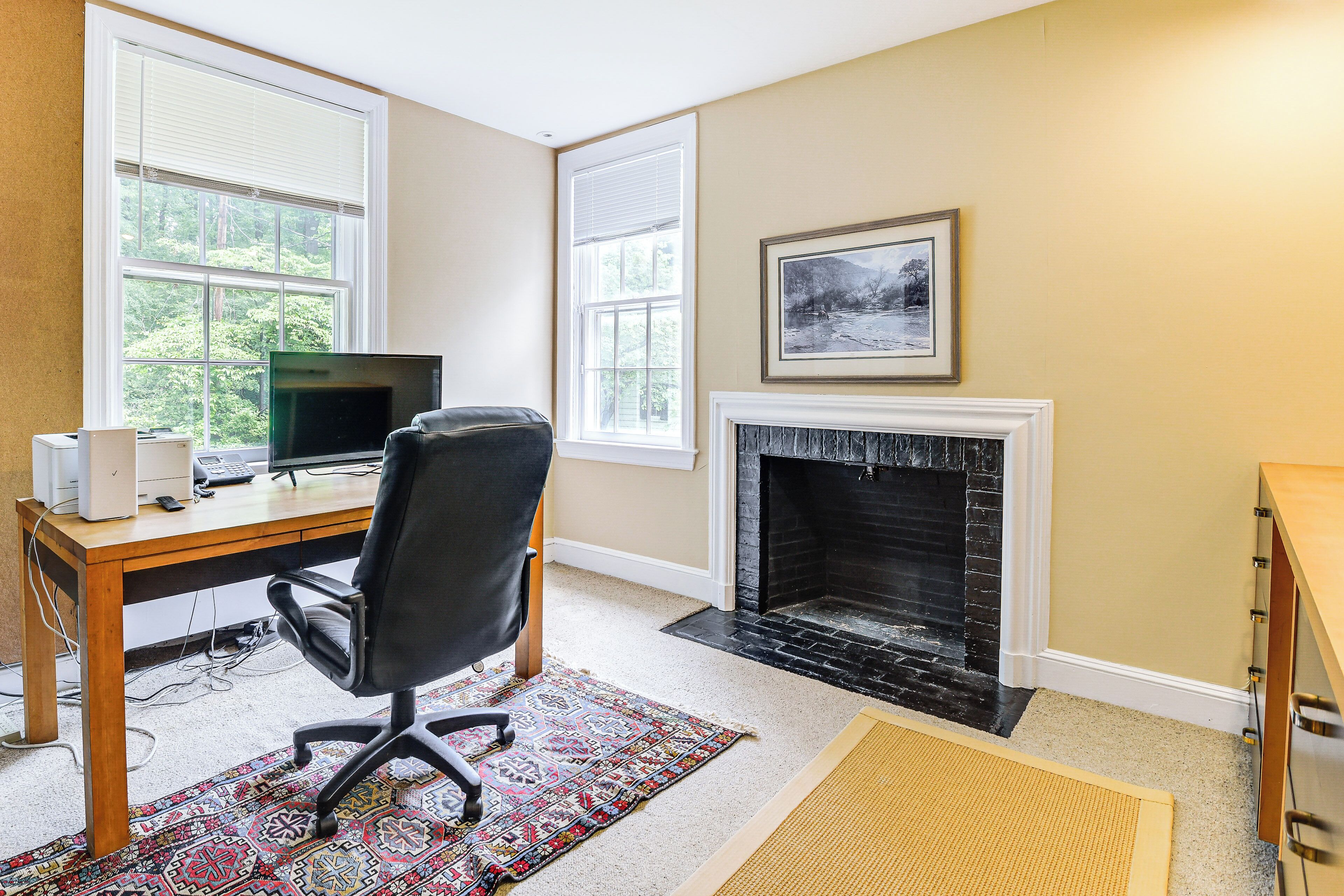 A room with a wooden desk, a black office chair, a fireplace, and a large window with blinds.