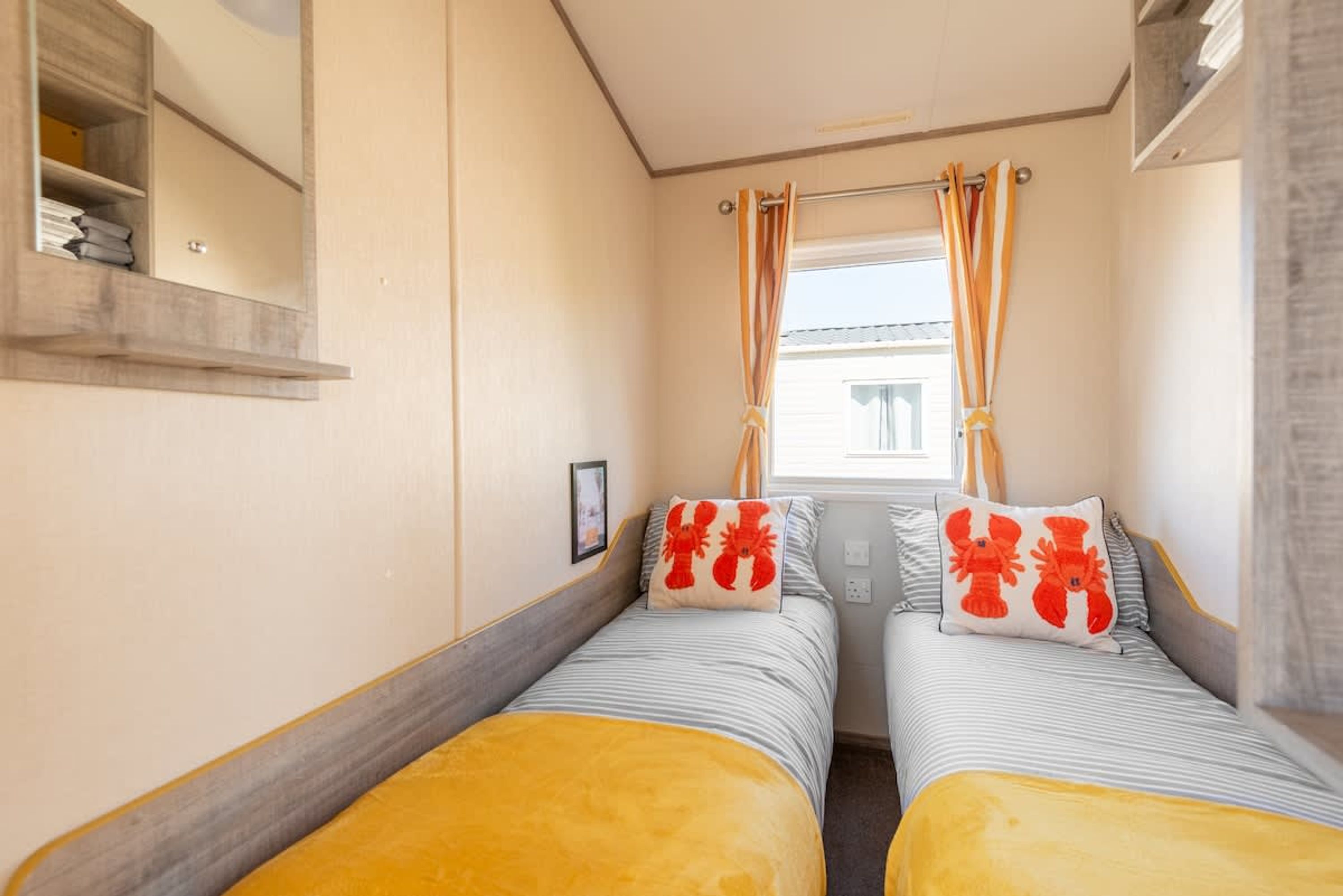 Une chambre de taille modeste, équipée de deux lits, chacun recouvert d’une couvre-lit à rayures et agrémenté d’un coussin orné de motifs représentant des homards rouges. On y trouve une fenêtre avec des rideaux, une petite étagère avec quelques objets et un tableau encadré accroché au mur.