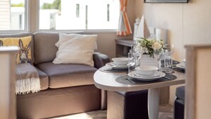 A dining area with a round table set for four, a sofa with cushions, and a window with curtains. - 3 Bedroom Caravan-seton Sands (Prestonpans)