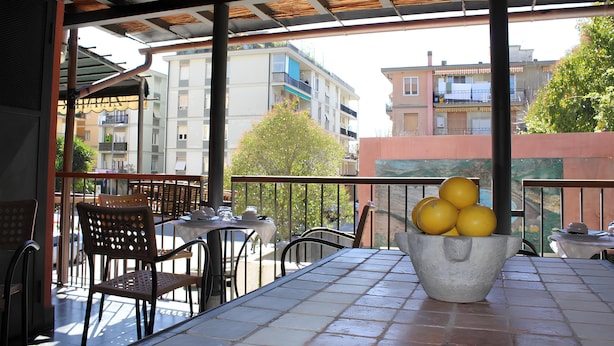 Un espace extérieur aménagé avec des tables et des chaises, un récipient en béton rempli d’oranges, et une vue sur des bâtiments et un arbre.