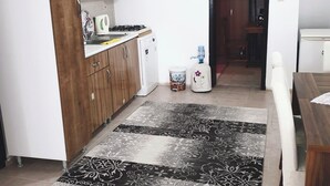 A compact kitchen with wooden cabinets, a small refrigerator, and a gas stove. There is a dining area with a table and chairs, and a patterned rug on the floor.