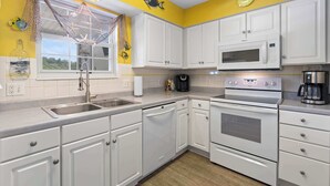Una cocina con armarios blancos, microondas, horno, fregadero y ventana con mosquitero.
