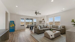 A spacious living room with a large gray sectional sofa, a coffee table, and a ceiling fan. There are large windows offering a view of the desert landscape.