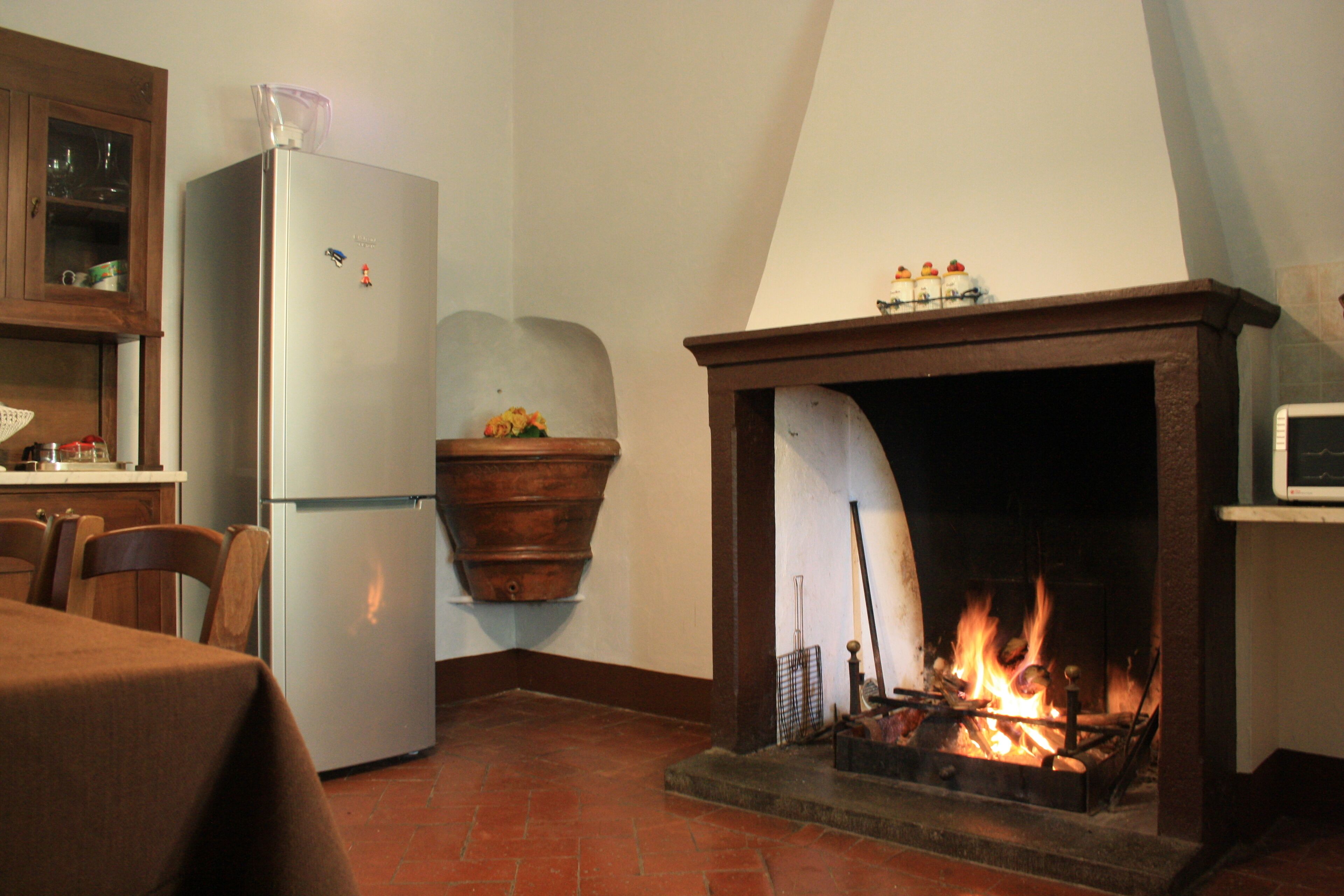 A kitchen with a refrigerator, a wooden cabinet, a dining table, and a fireplace with a fire.