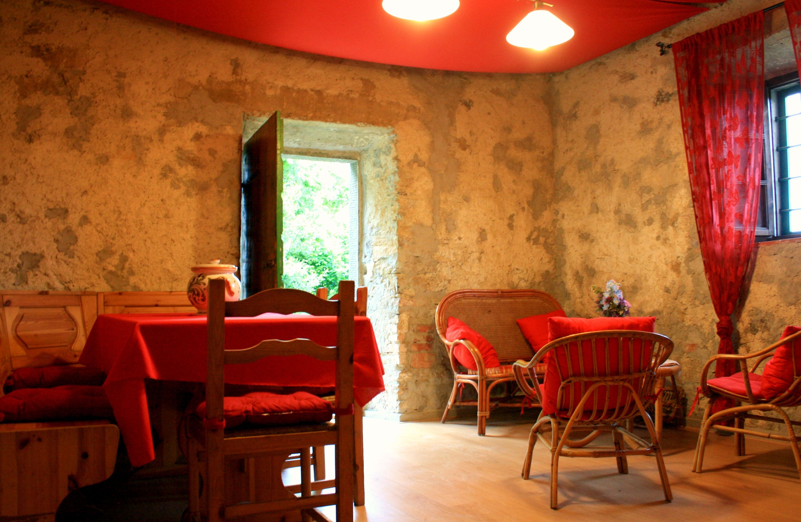 Une pièce rustique avec une table et des chaises en bois, un canapé en osier et une fenêtre ornée de rideaux rouges.