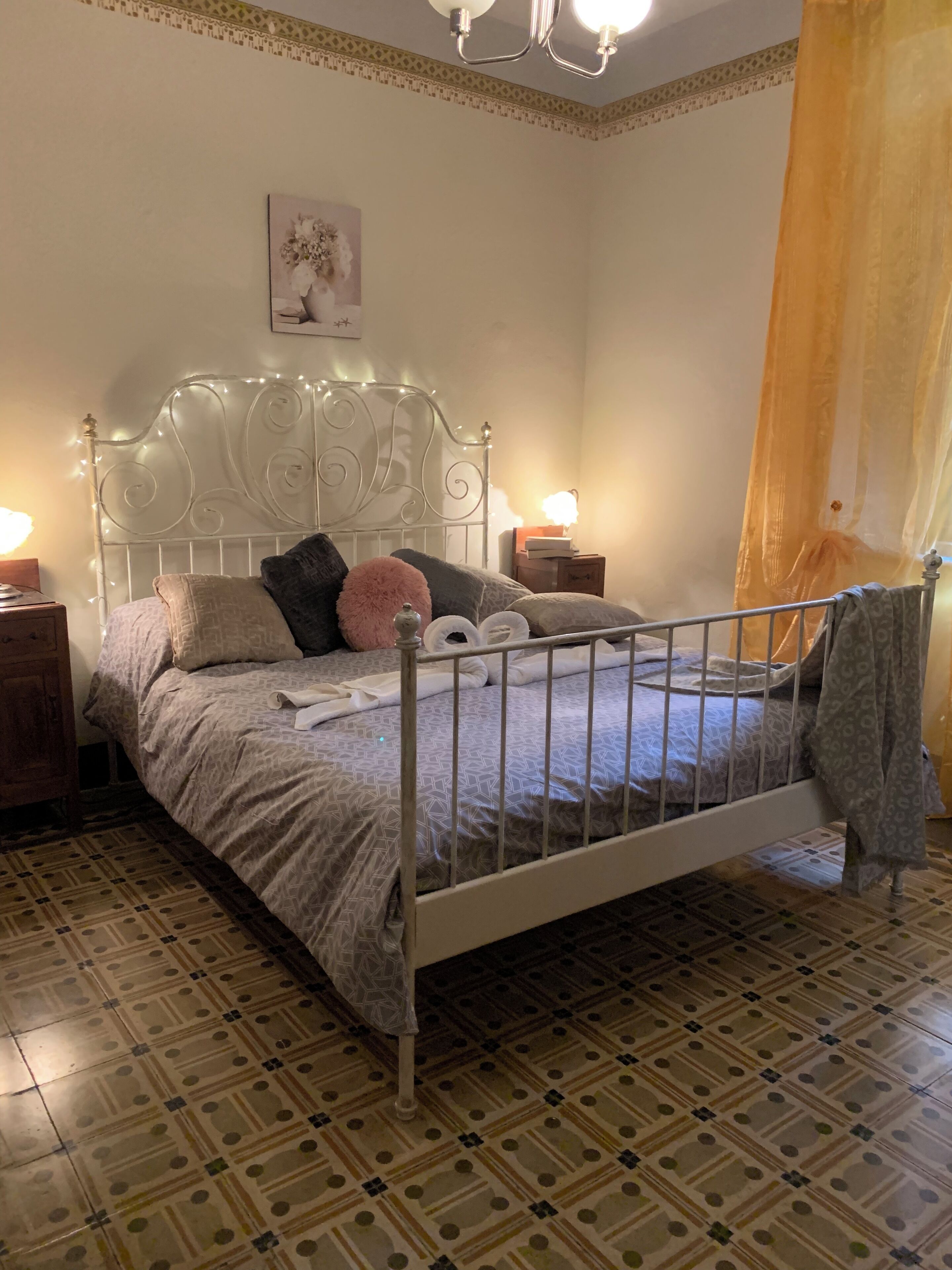 Ein ordentlich bezogenes Bett mit Metallgestell, zwei Nachttische mit Lampen, geflieste Böden mit Muster und ein dekorativer Kopfteil.