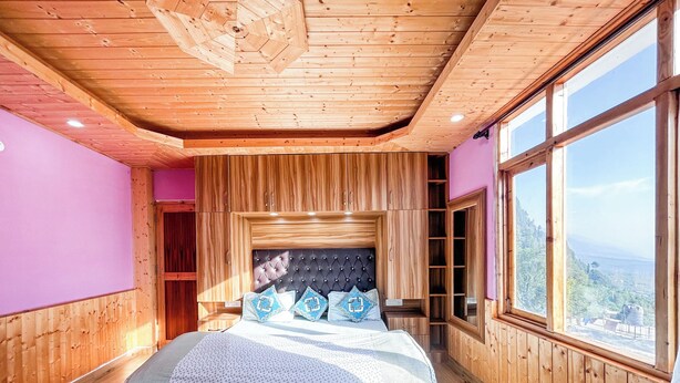 A bedroom with a wooden ceiling, a large bed with patterned bedding, a headboard, and a view through a large window. - MonAmis Home (Manali)