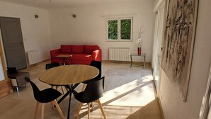 Une pièce avec un canapé rouge, une table ronde en bois et des chaises noires, un petit radiateur blanc et une fenêtre donnant sur un paysage verdoyant.