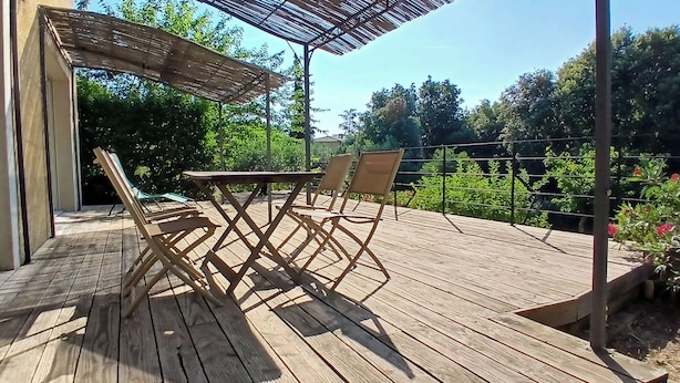 Une terrasse en bois avec une table et des chaises, entourée d’une clôture en métal et de verdure.