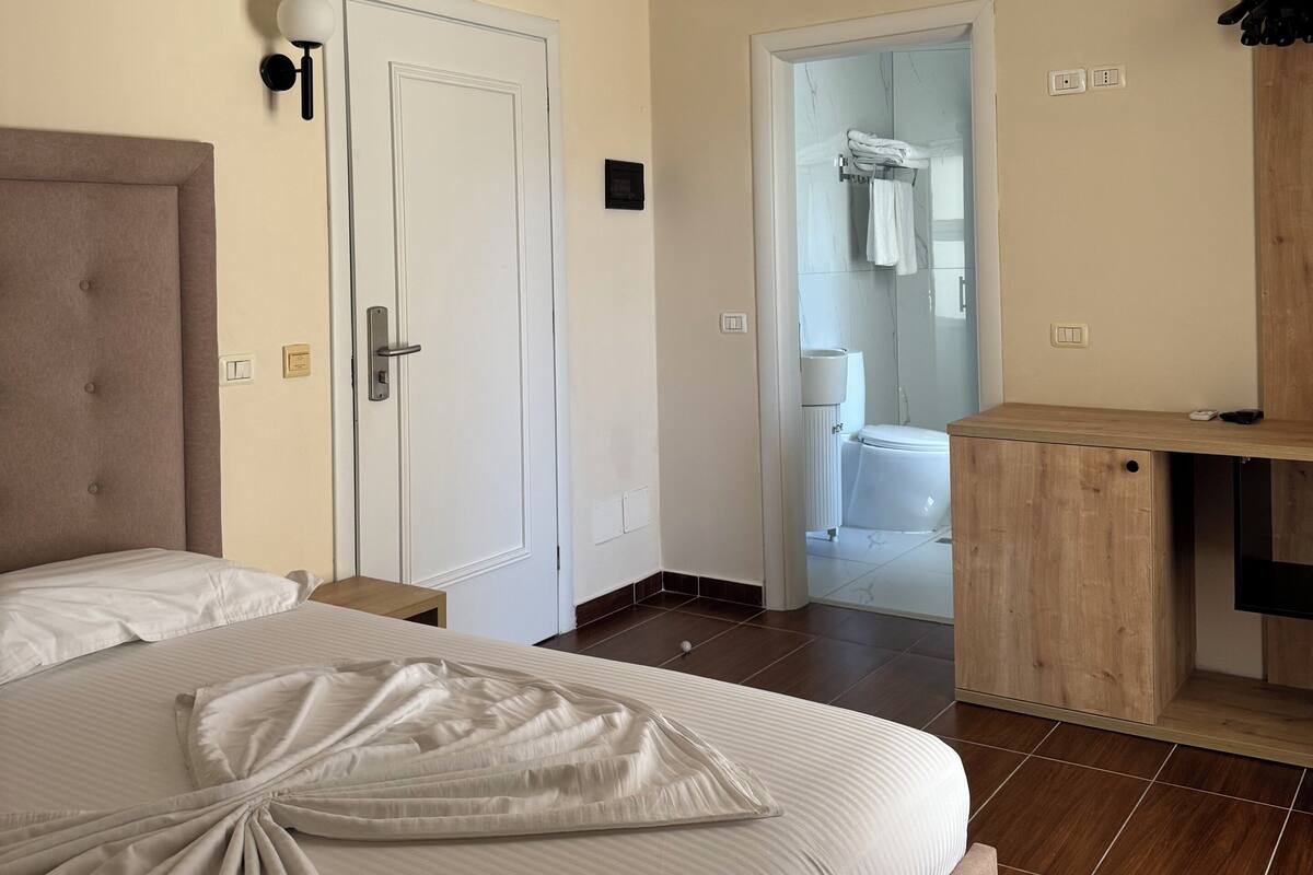 Una camera d'albergo con un letto, una scrivania in legno, un bagno parzialmente visibile attraverso una porta aperta e un lampadario fissato alla parete.