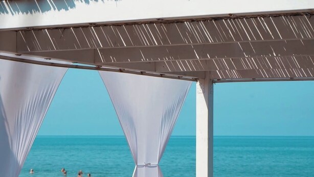 Un área de descanso frente a la playa con vista al mar, que incluye sillones de madera, una mesa pequeña y un toldo blanco.