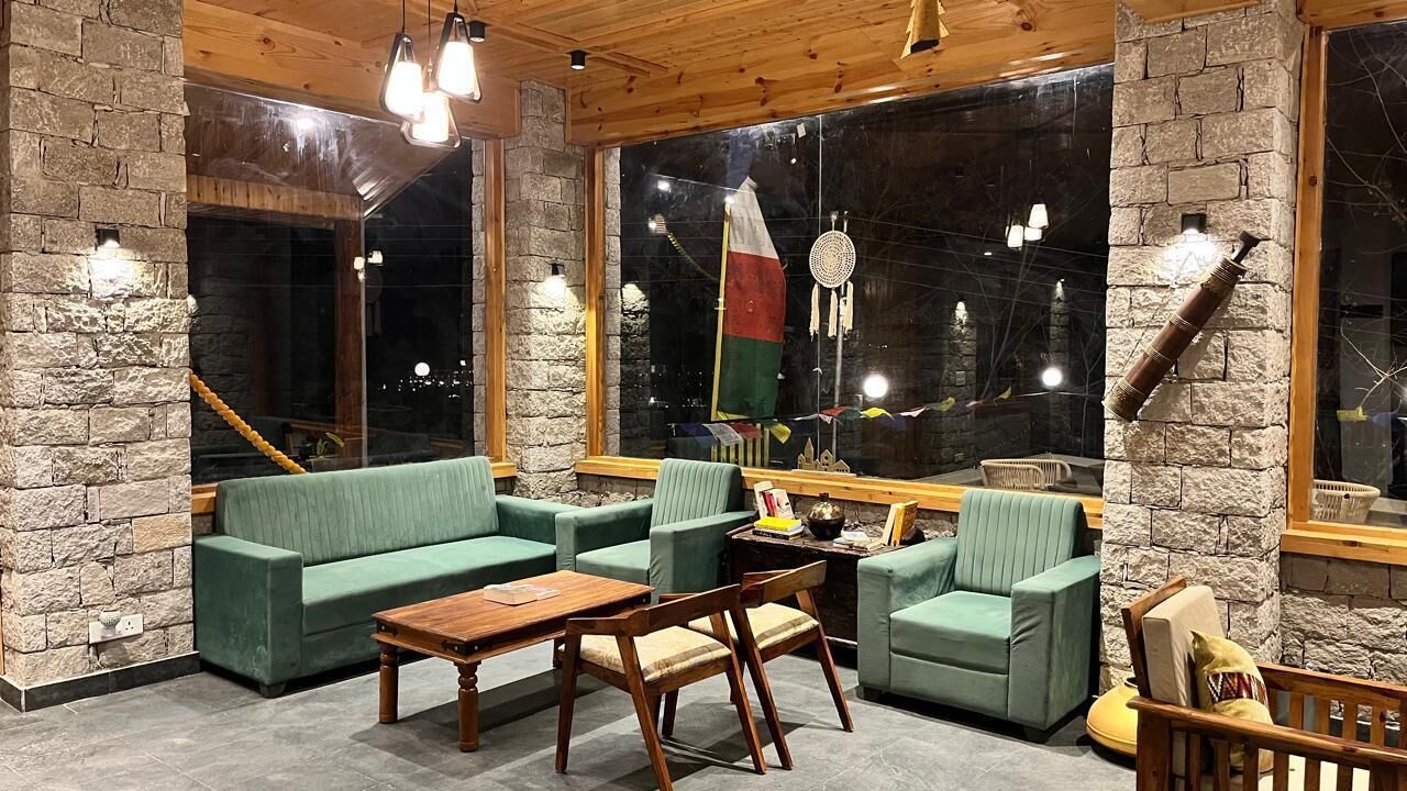 Ein gemütlicher Raum mit Steinwänden, grünen Sofas, einem Holztisch und einem großen Fenster mit Flagge und Traumfänger.