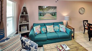 Ein Wohnzimmer mit einem türkisfarbenen Sofa, Kissen mit Seestern-Muster, einem Couchtisch aus Holz und einem Strand-Themenbild an der Wand.