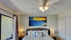 Une chambre d’hôtel avec un lit, un tableau représentant une scène de plage, un ventilateur de plafond et une vue sur le balcon extérieur.