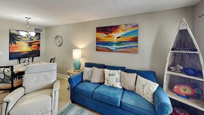 Un salon avec un canapé bleu, un fauteuil blanc, une table à manger avec des chaises, une horloge et deux tableaux.