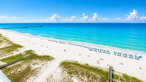 A beach with a long pier extending into the ocean, a sandy beach with rows of blue umbrellas, and a clear blue sky. - Phoenix Gulf Shores Ii 1203 3 Bedroom Condo (Gulf Shores)