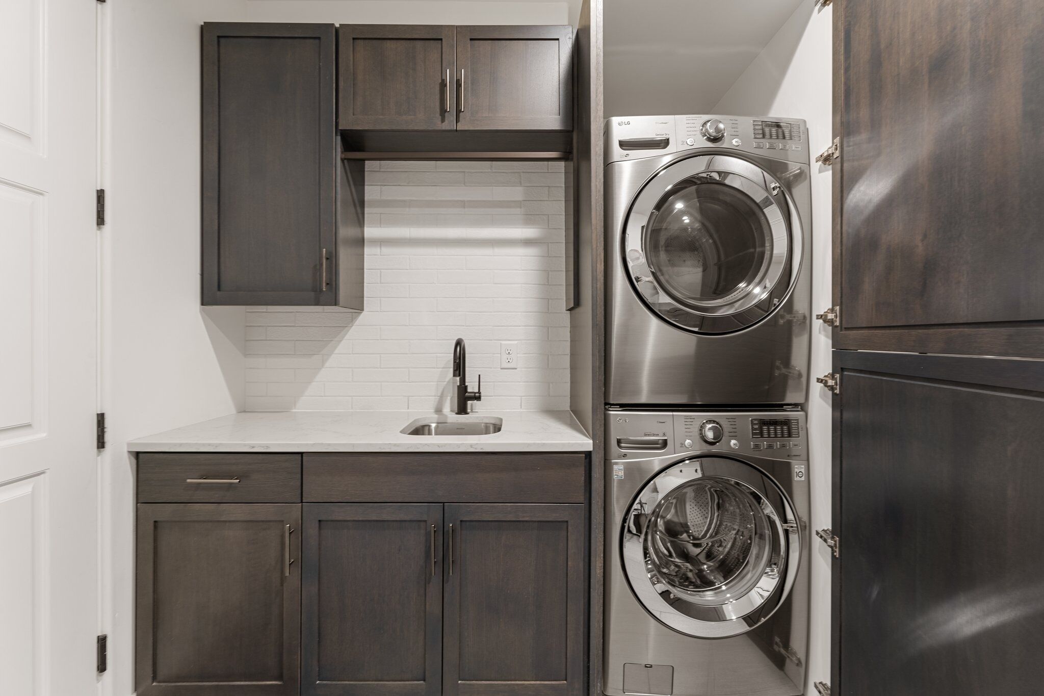 A compact laundry room with a built-in washer and dryer, a sink, and dark wood cabinets.