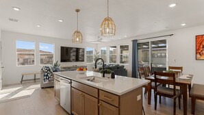 Una cocina moderna con una isla central, luces colgantes y una zona de comedor con sillas y mesa de madera.
