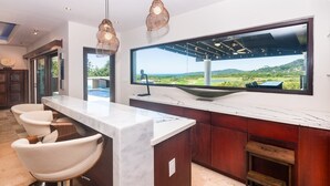 A modern kitchen with a marble countertop, two pendant lights, a large window with a view, and a bar area with stools. - Pristine Bay 1005- Villa Loce 4 Bedroom Home (Roatan)