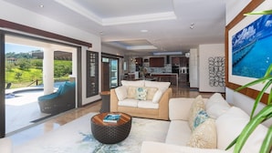 A spacious living room with a large window, a sofa set, a coffee table, and a kitchen in the background. - Pristine Bay 1005- Villa Loce 4 Bedroom Home (Roatan)