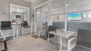 A modern indoor seating area with metal tables and chairs, large windows, and a television screen displaying a beach scene.