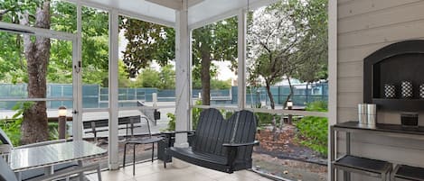 A covered patio area with a swing chair, a table, and chairs, overlooking a garden and a building in the background.