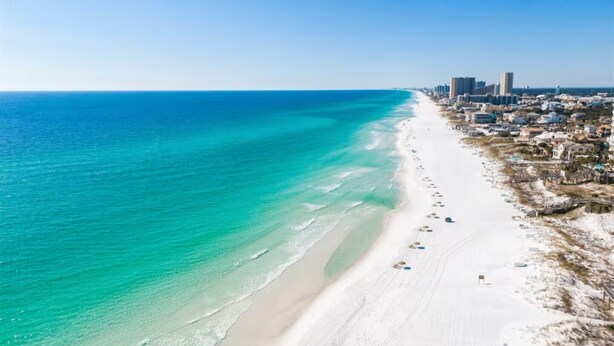 A coastal cityscape with a wide beach, clear blue waters, and a dense urban area with high-rise buildings.