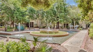 A landscaped garden with a central fountain, surrounded by trees and a paved walkway.