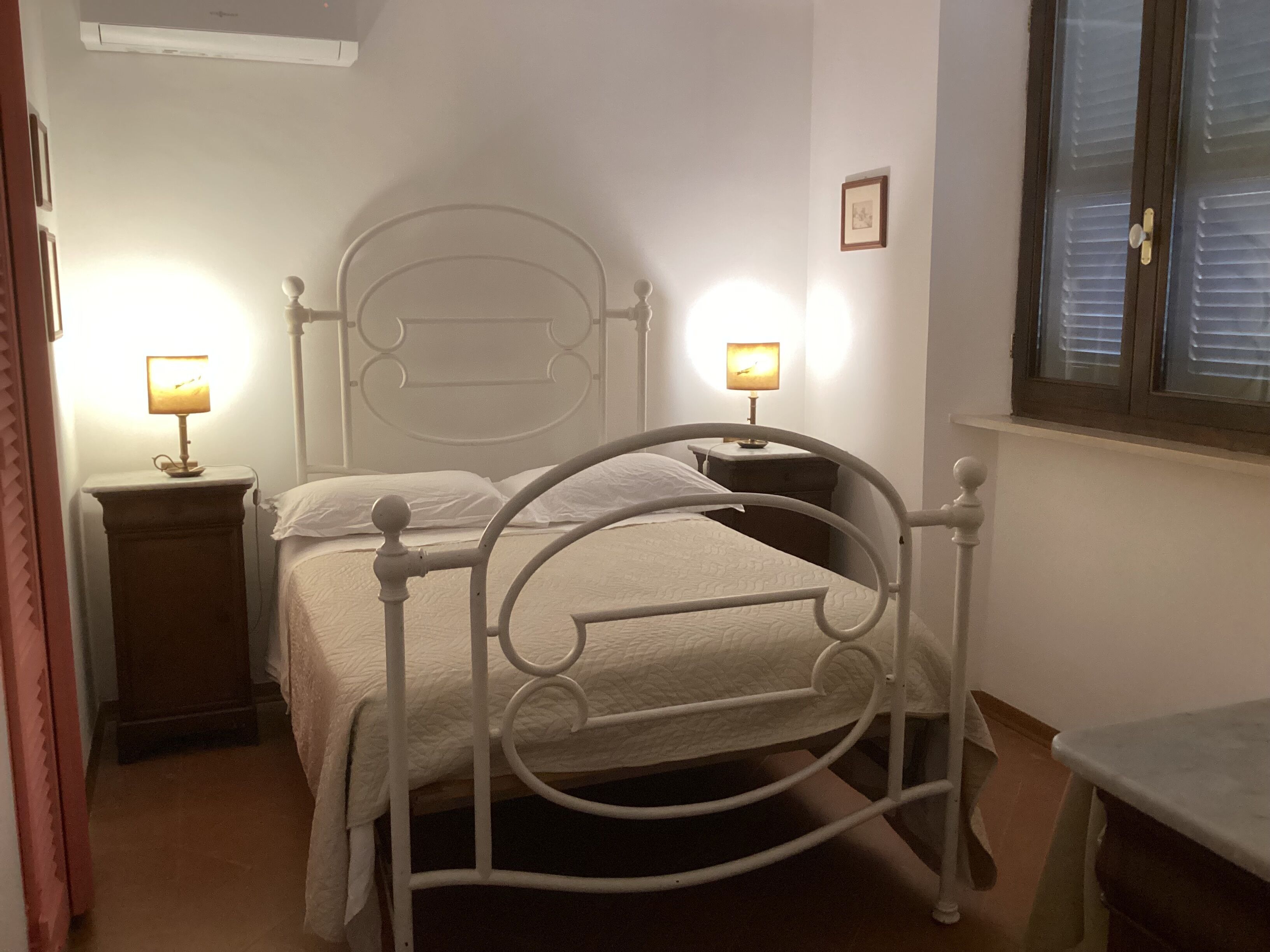 A white metal bed with a headboard, two bedside tables with lamps, a window with blinds, and a small framed picture on the wall.