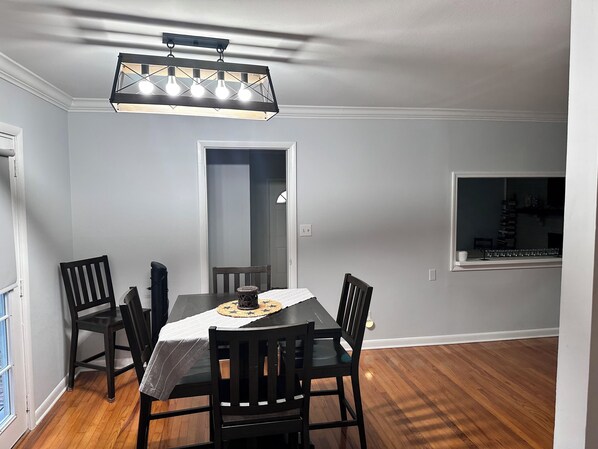 Un comedor con una mesa rectangular, seis sillas, una araña y una ventana que da a una estantería.