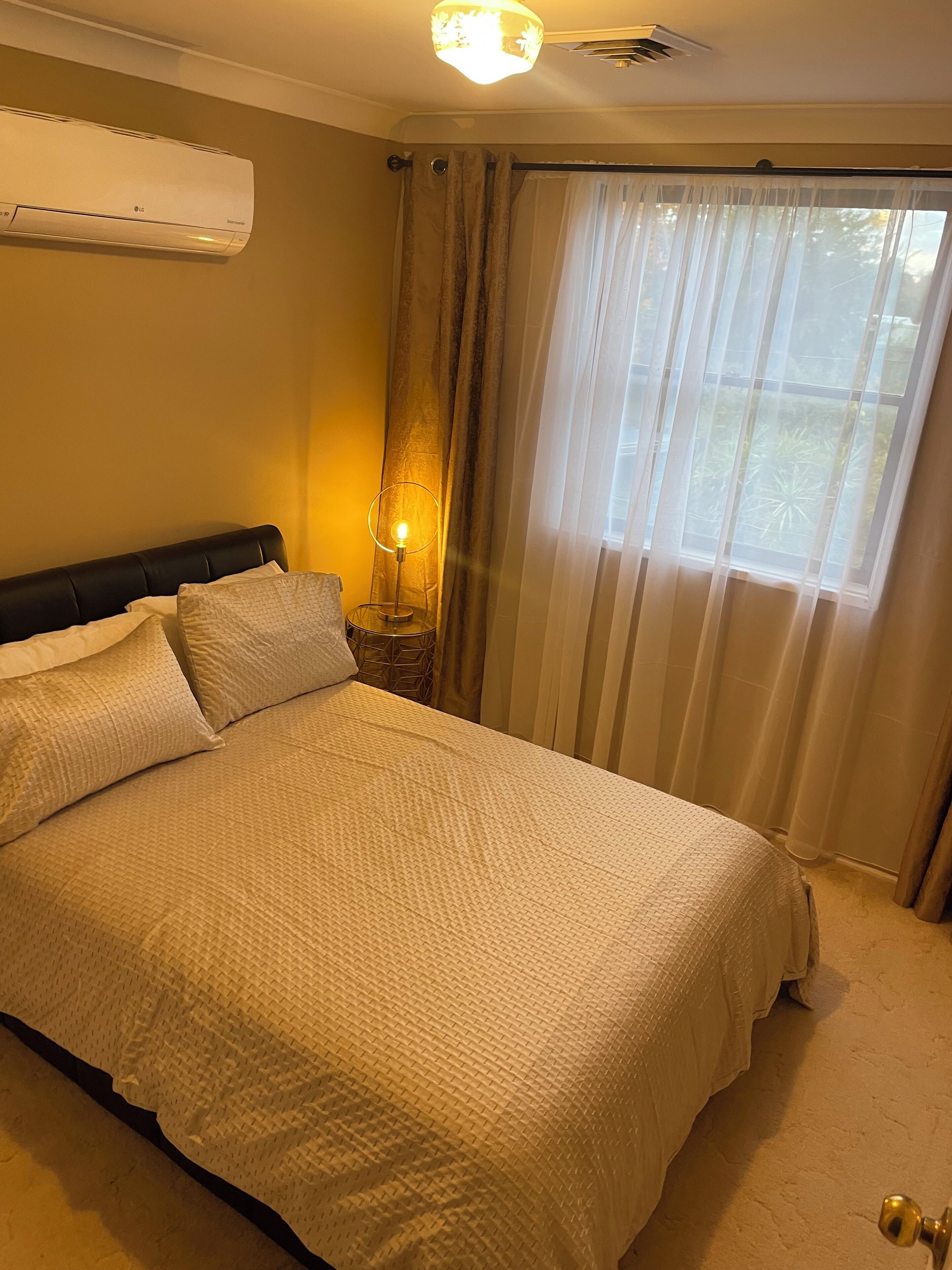 Un lit bien fait, recouvert d’une couette blanche à texture intéressante, une lampe de chevet et une fenêtre ornée de rideaux légers.
