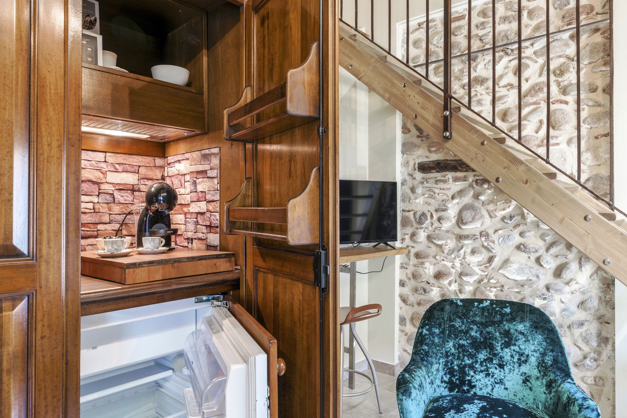 A compact kitchen with a coffee machine, a stone wall, and a wooden staircase.