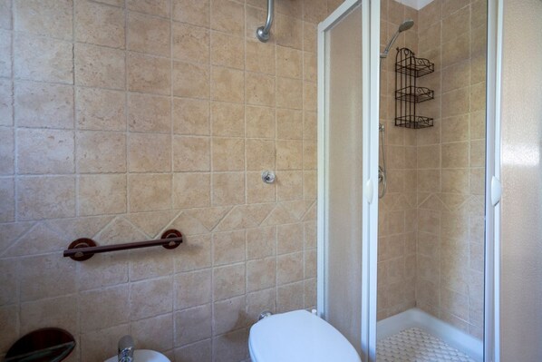 A bathroom with beige tiles, a white toilet, a shower with a metal rack, and a grab bar.