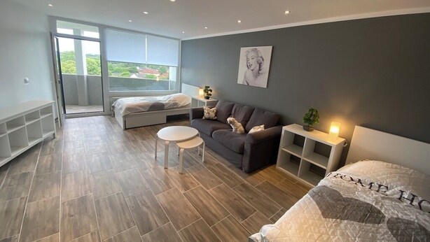 A modern bedroom with a large bed, a sofa, a small table, and a nightstand. There is a window with a view of greenery and buildings.