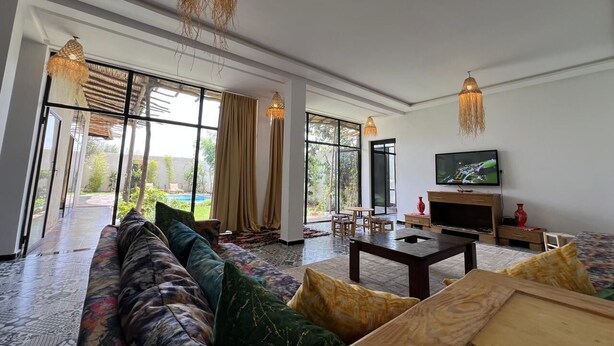 A spacious living room with a large sofa, a coffee table, and a television. There are large windows providing a view of the outdoors. - Family villa, private pool, not overlooked (Marrakech)