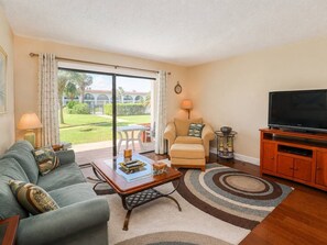 A living room with a sofa, armchair, coffee table, TV stand, and a view of a garden through a sliding door. - Walk to St. Augustine Beach | Poolside Patio (St. Augustine)
