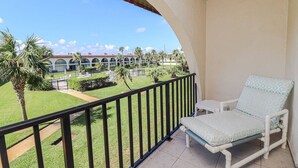 Un balcon avec une chaise blanche et une vue sur un complexe hôtelier bordé de palmiers et sur un bâtiment doté d’arcades.