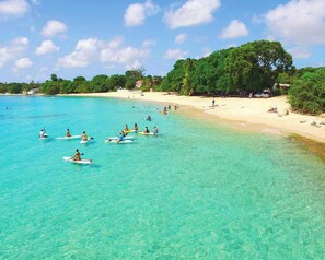 A beach with clear turquoise water, people paddleboarding, and a sandy shore with trees. - New CHIC 3BR Villa, Walk to Beach! Pool & Terrace (Lower Carlton)