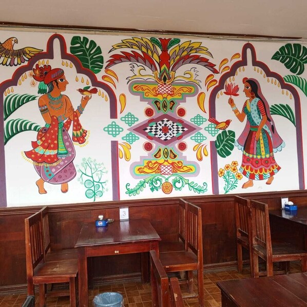 A restaurant interior with wooden tables and chairs, a mural with cultural artwork, and a tiled floor.
