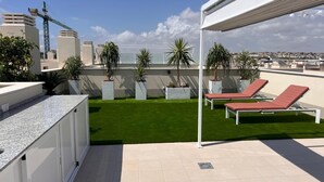 Una terraza en la azotea con césped verde, dos sillones rojos y plantas en macetas.