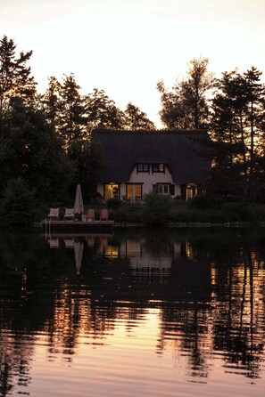 A house with a thatched roof, a pool, and a reflection in the water.