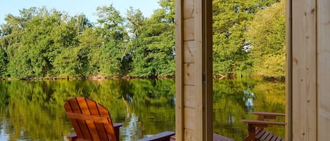 Een houten terras met een houten stoel en tafel, met uitzicht op een kalm meer met bomen op de achtergrond.