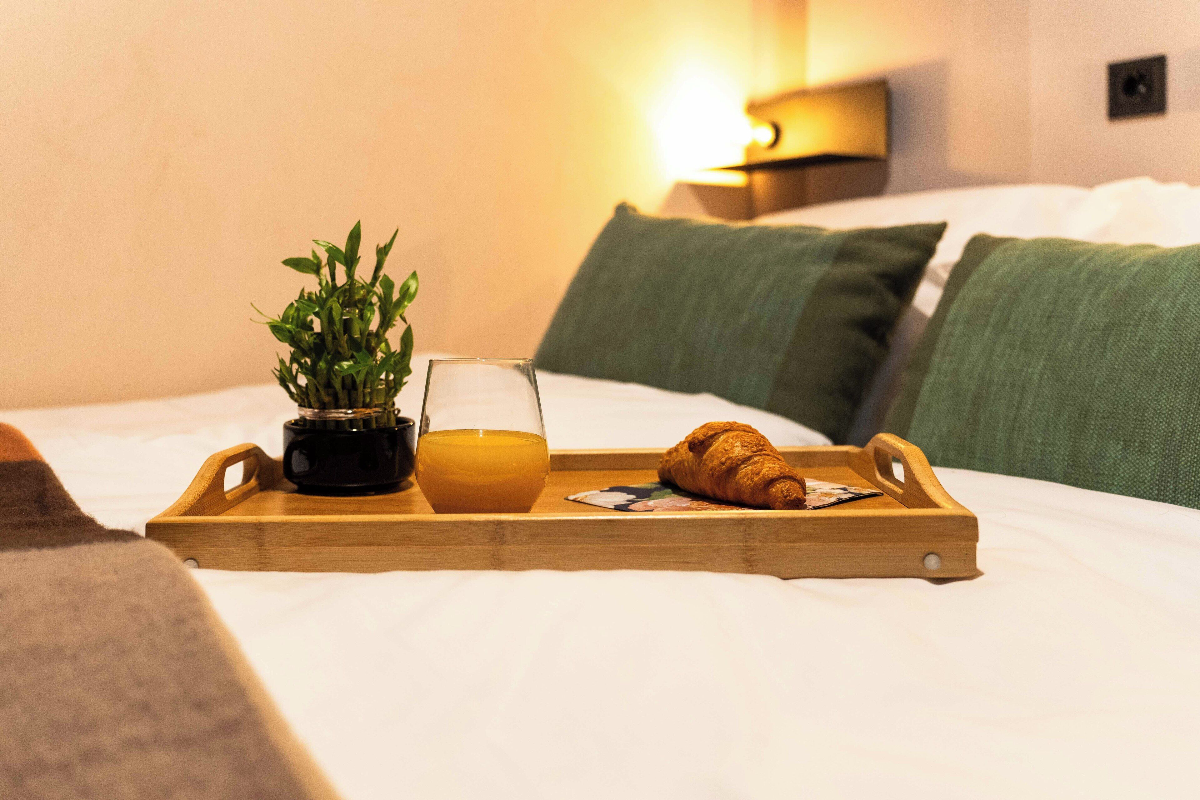Una bandeja de madera con una planta, un vaso de jugo de naranja y un croissant sobre una cama con almohadas verdes y una lámpara fijada a la pared.
