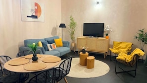 Sala de estar com sofá, televisão, mesa de jantar, cadeiras e plantas decorativas.