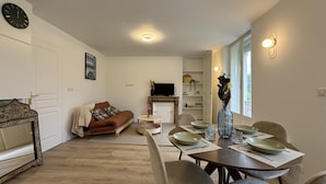 En moderne stue med sofa, spisebord og stole. Der er pejs, fjernsyn og et ur på væggen.