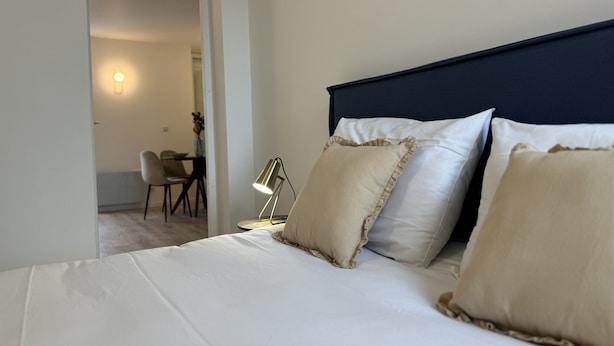 En pænt redt seng med hvide og beige puder, en mørk sengegavl og en skrivebordslampe i et rum med en dør, der fører til et andet område.